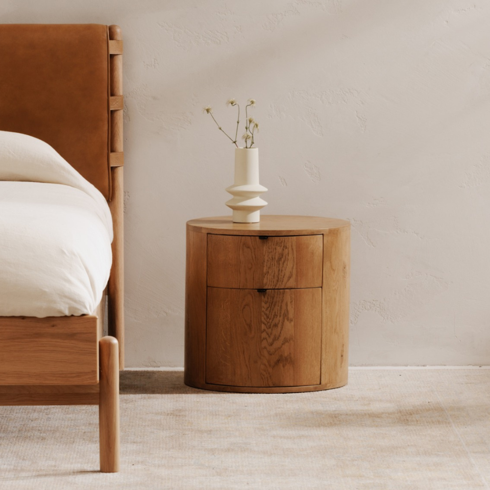 Solid oak Theo nightstand with two drawers, featuring a curved design and minimalist style, placed beside a bed.