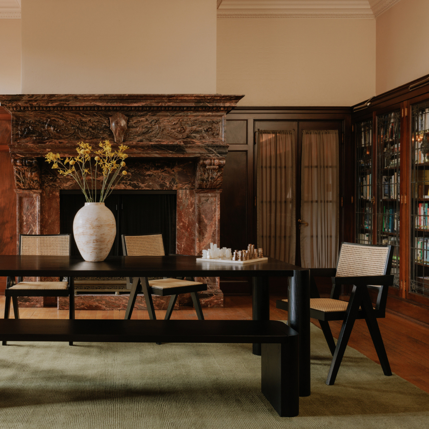 Takashi chair with  black elm wood frame and natural rattan seat, set in a classic room with fireplace, dining table, and vase with yellow flowers.