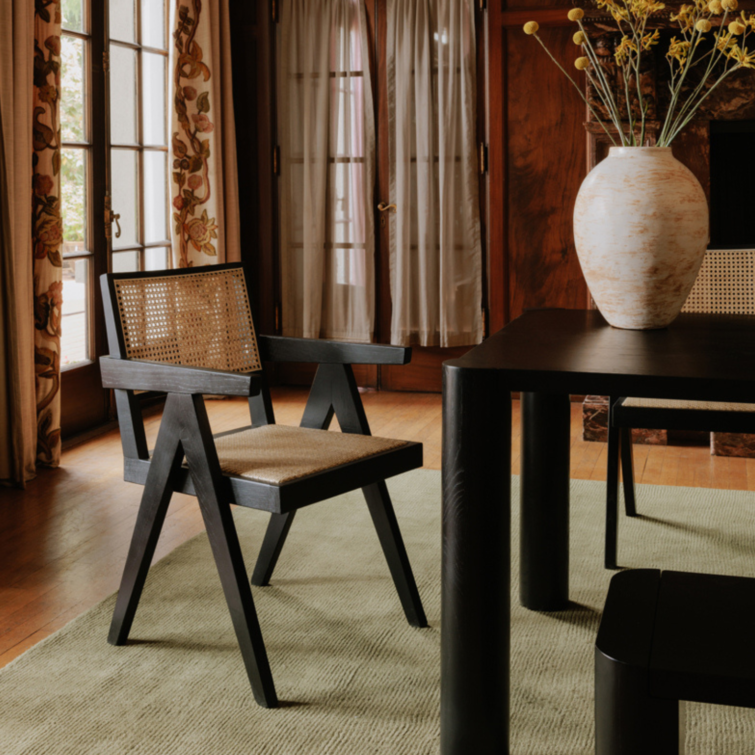 Black Takashi chair with rattan seat in an elegant room, next to a dark dining table with a large ceramic vase.